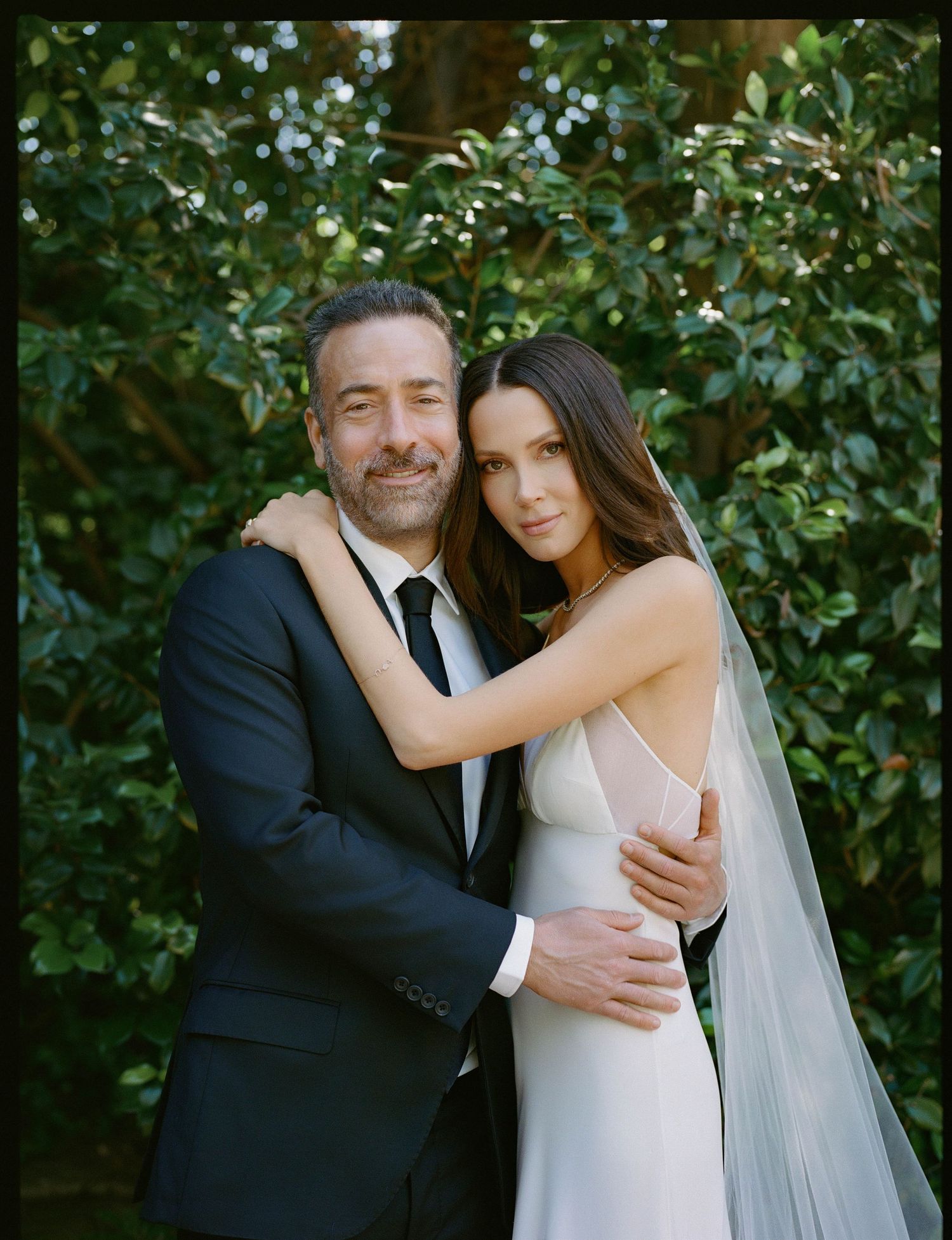 Couple in wedding attire embracing outdoors, groom in black suit, bride in white gown with veil, lush green foliage background.