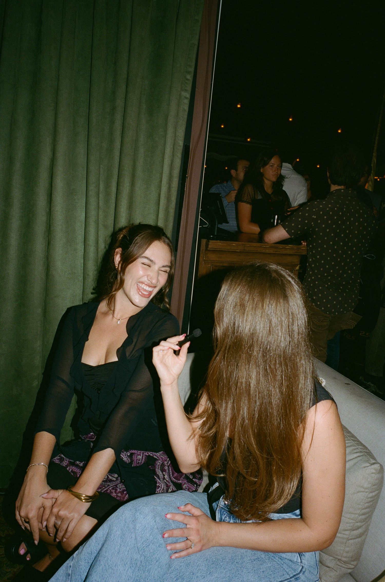Two young women laughing and chatting together at a dimly lit bar or nightclub venue.