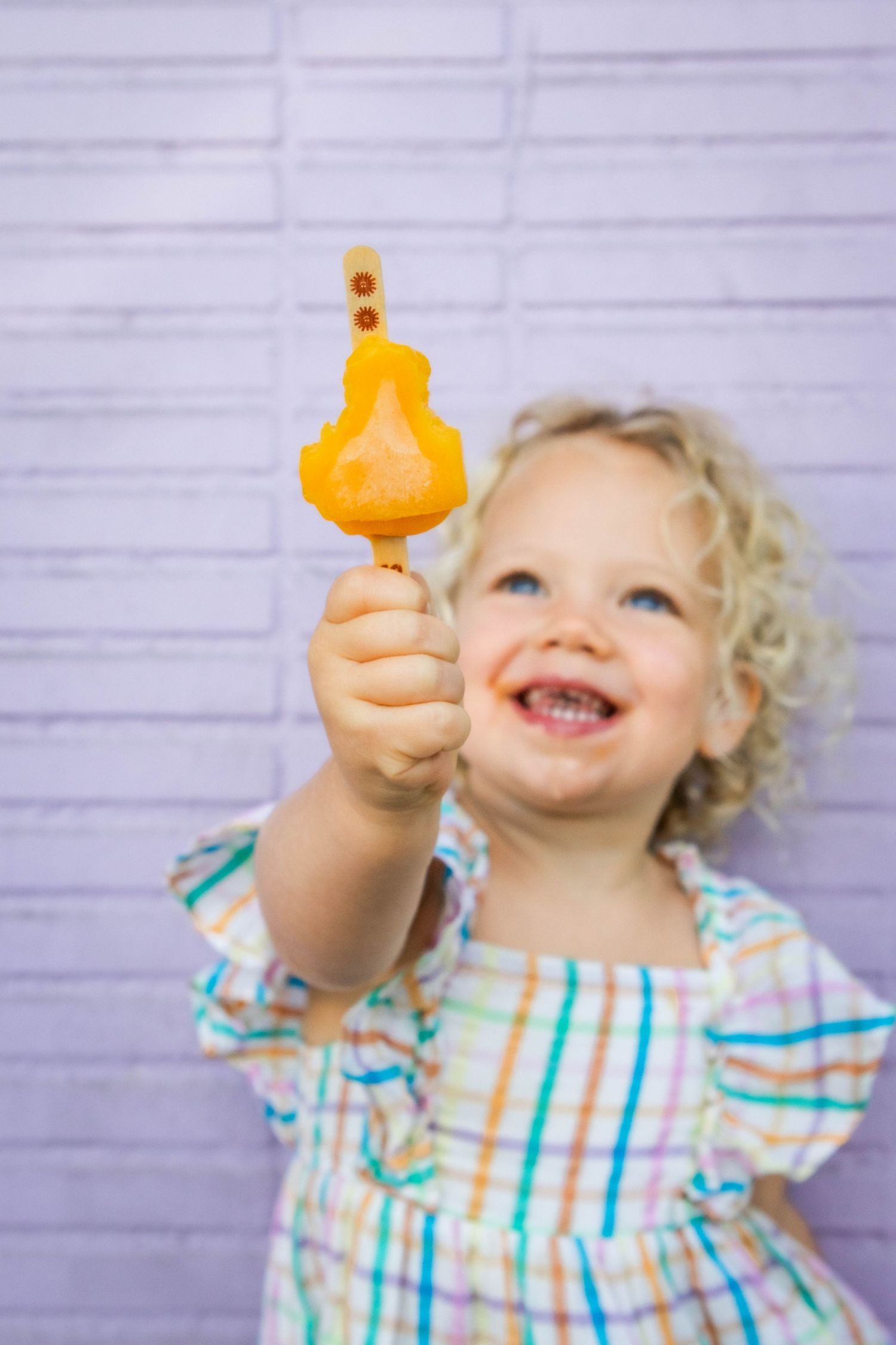 A happy child holding up a yellow duck-shaped popsicle against a purple wall.