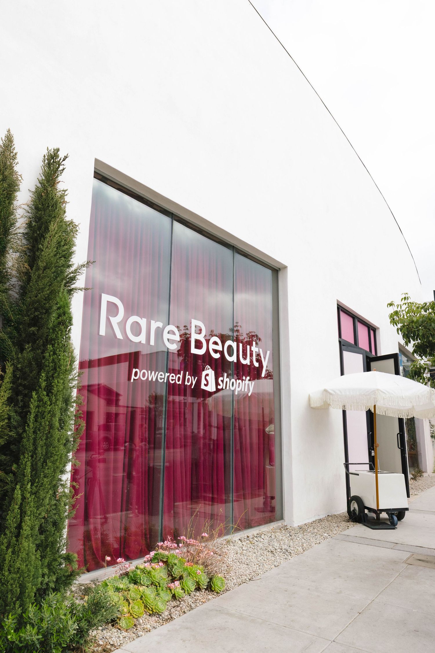 Modern storefront with 'Rare Beauty' signage and pink translucent panels, featuring cypress trees and white exterior walls.