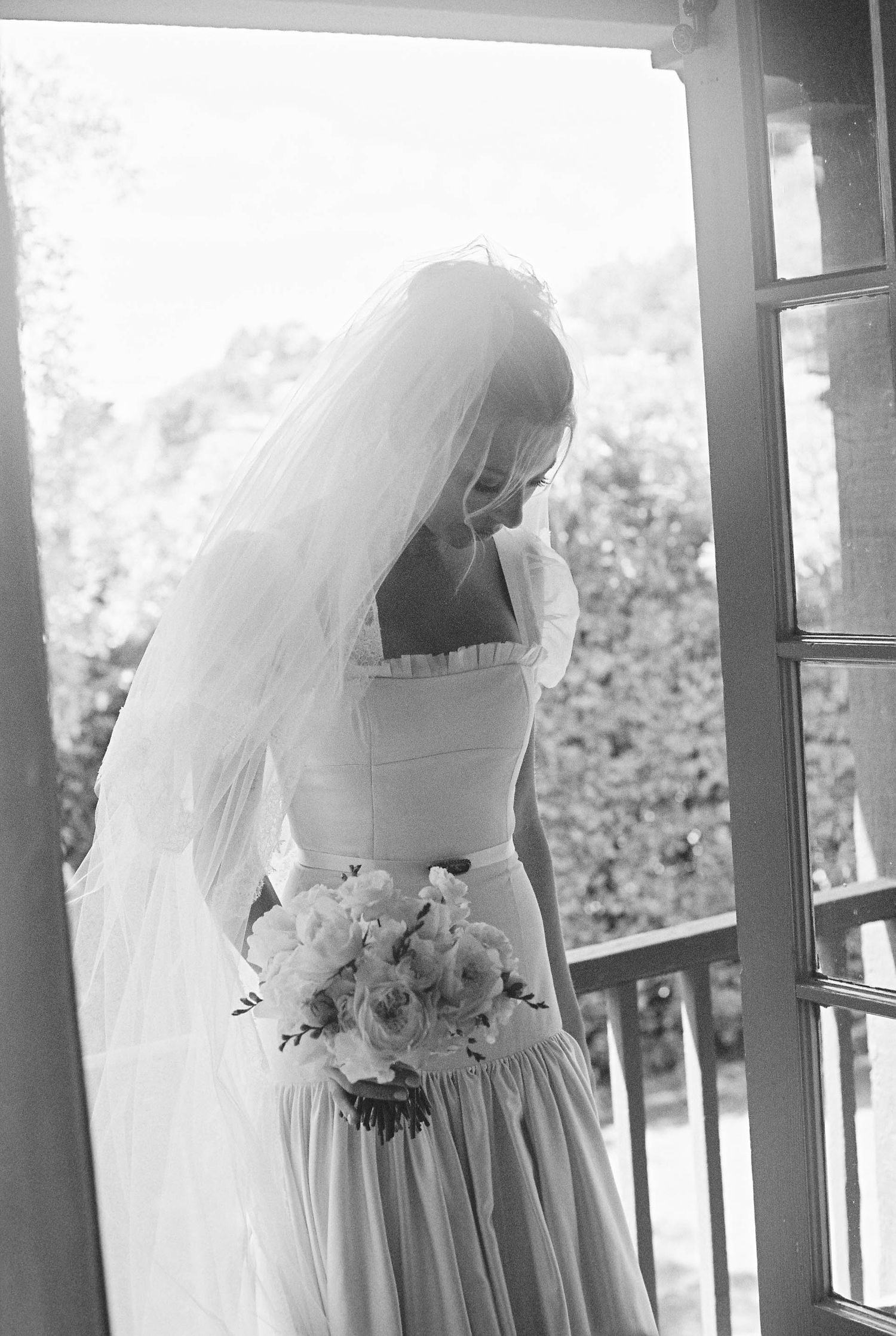 Black and white photo of bride in wedding dress holding bouquet on porch.