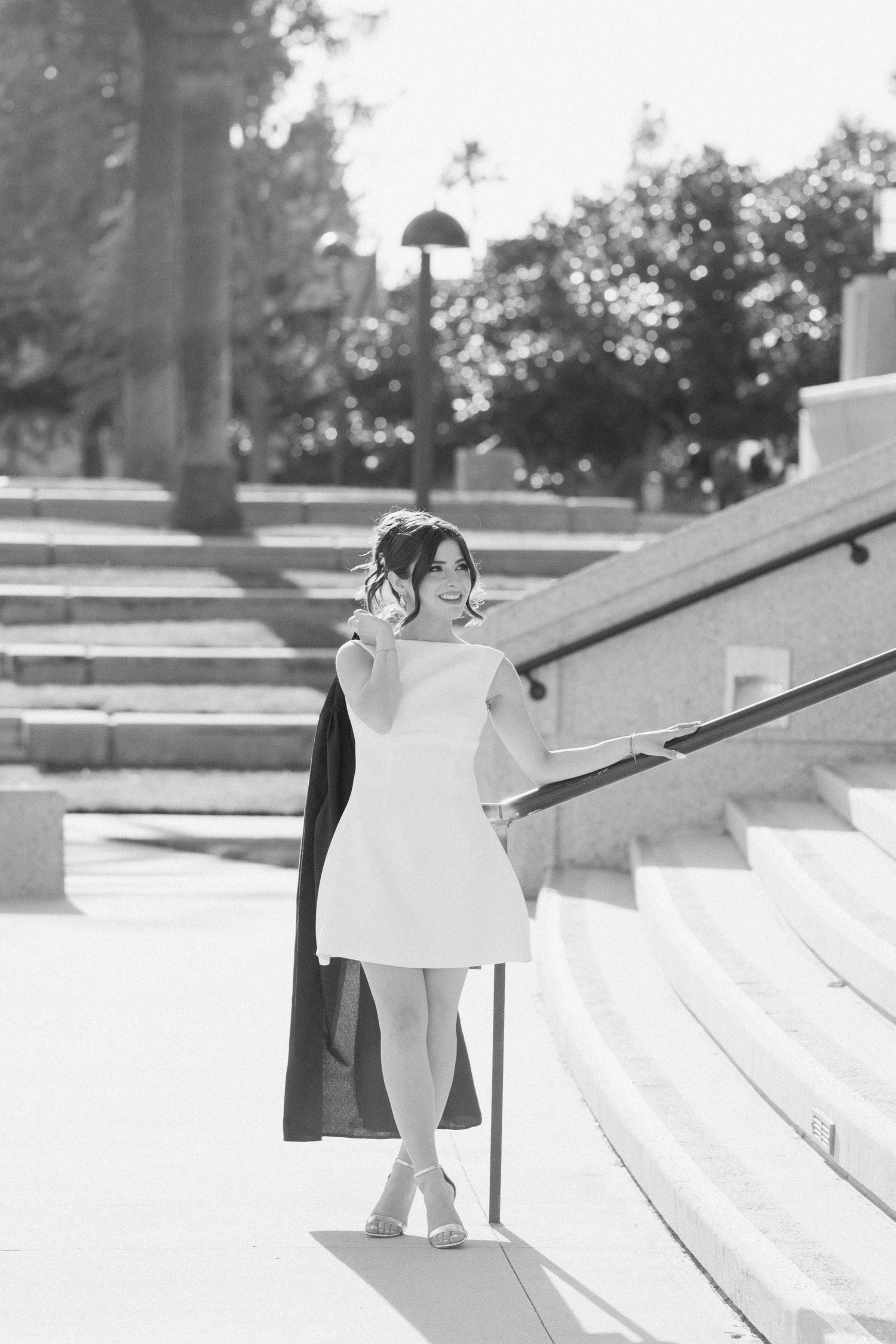 Graduate in white dress poses on stone steps in black and white photo.