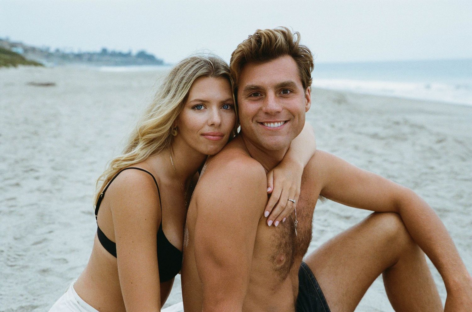 A couple embraces on a sandy beach at sunset with ocean waves in the background.
