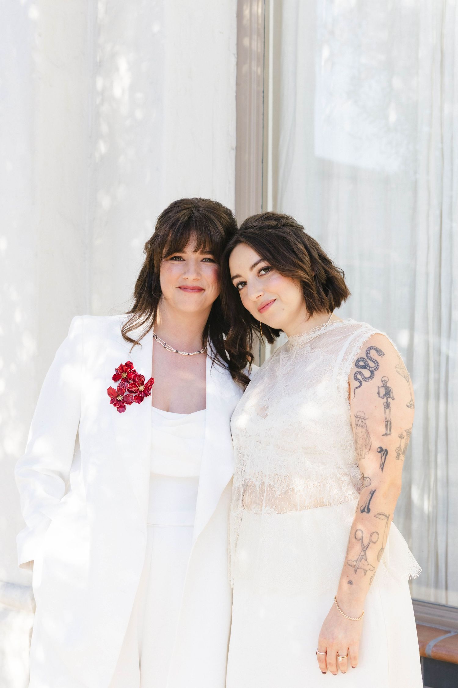 Two people in white outfits posing together in front of sheer curtains.