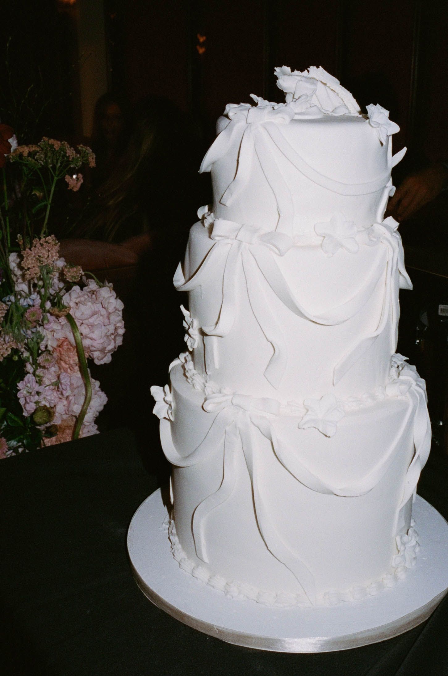 Elegant three-tier white wedding cake with fondant draping and butterfly decorations beside pink floral arrangement.