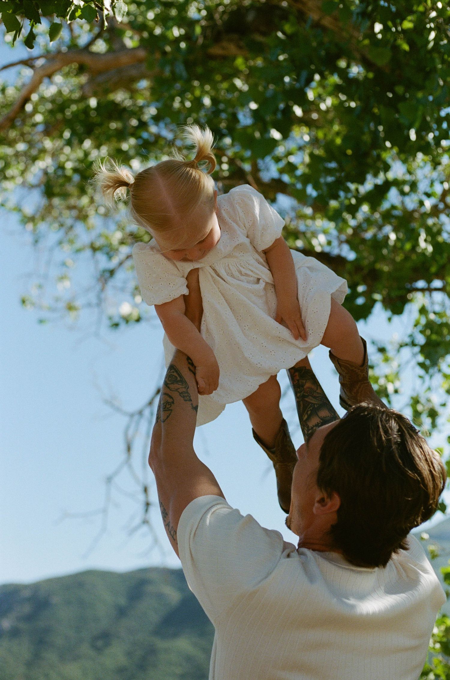 A tattooed parent joyfully lifts a smiling toddler girl in a white dress high in the air outdoors.