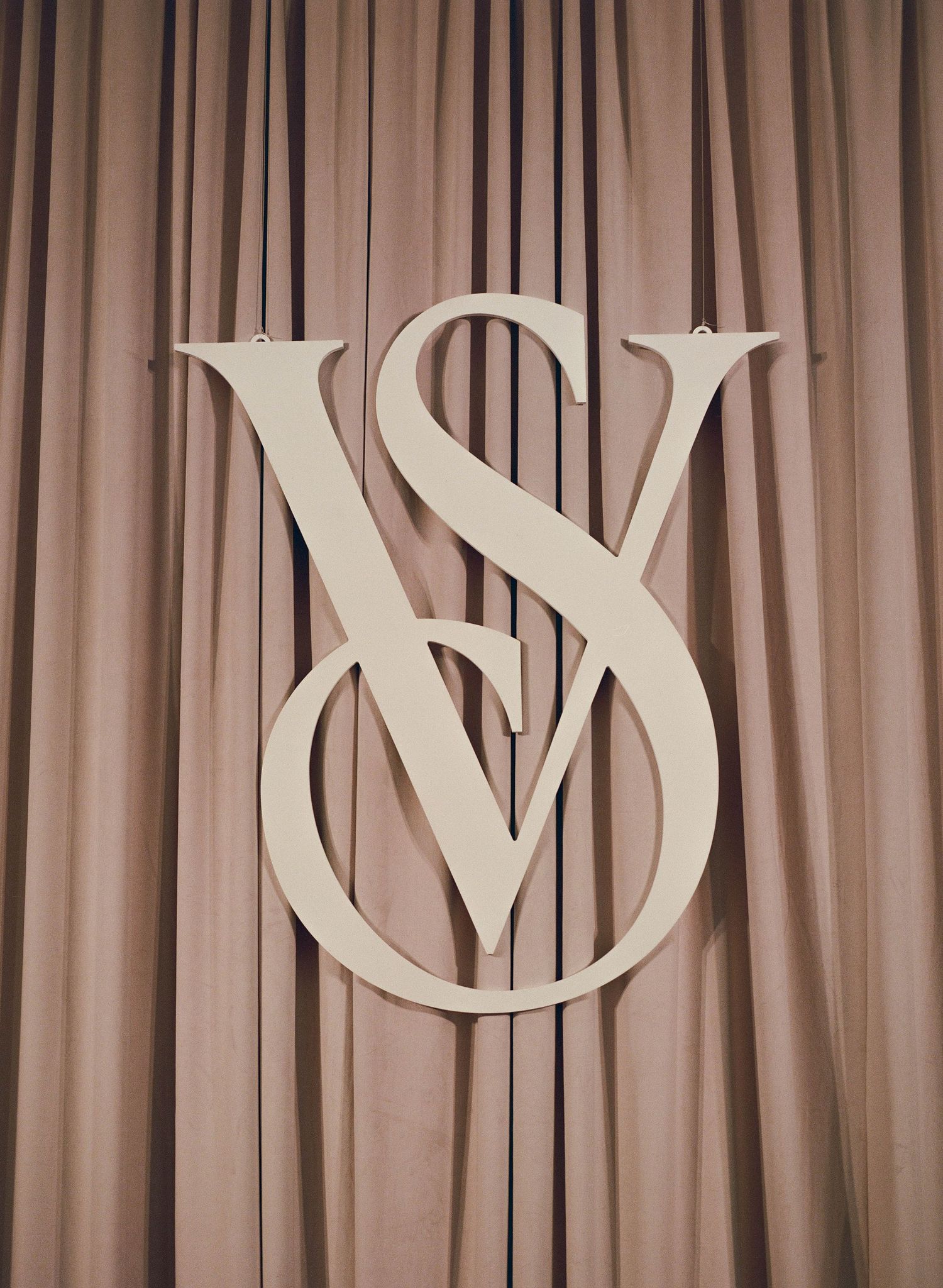 Cream interlocking V and S monogram sign hanging against soft blush pink draped curtains.