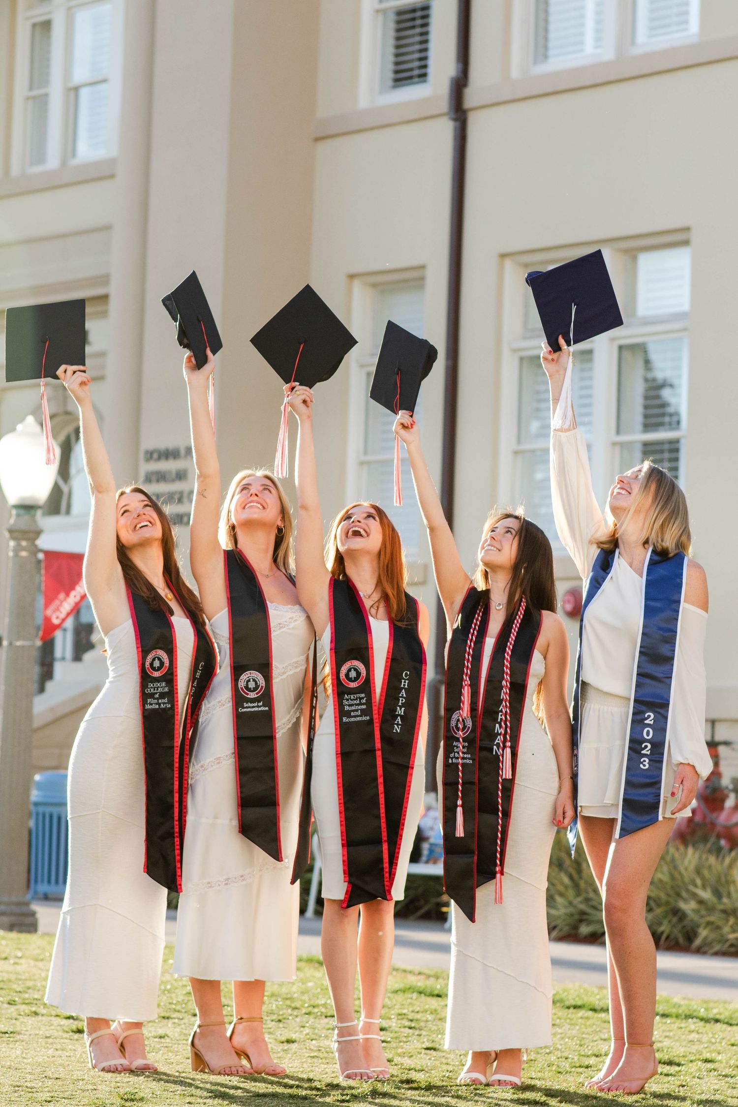 College graduates in white dresses and black graduation stoles toss their caps in celebration on a sunny day.