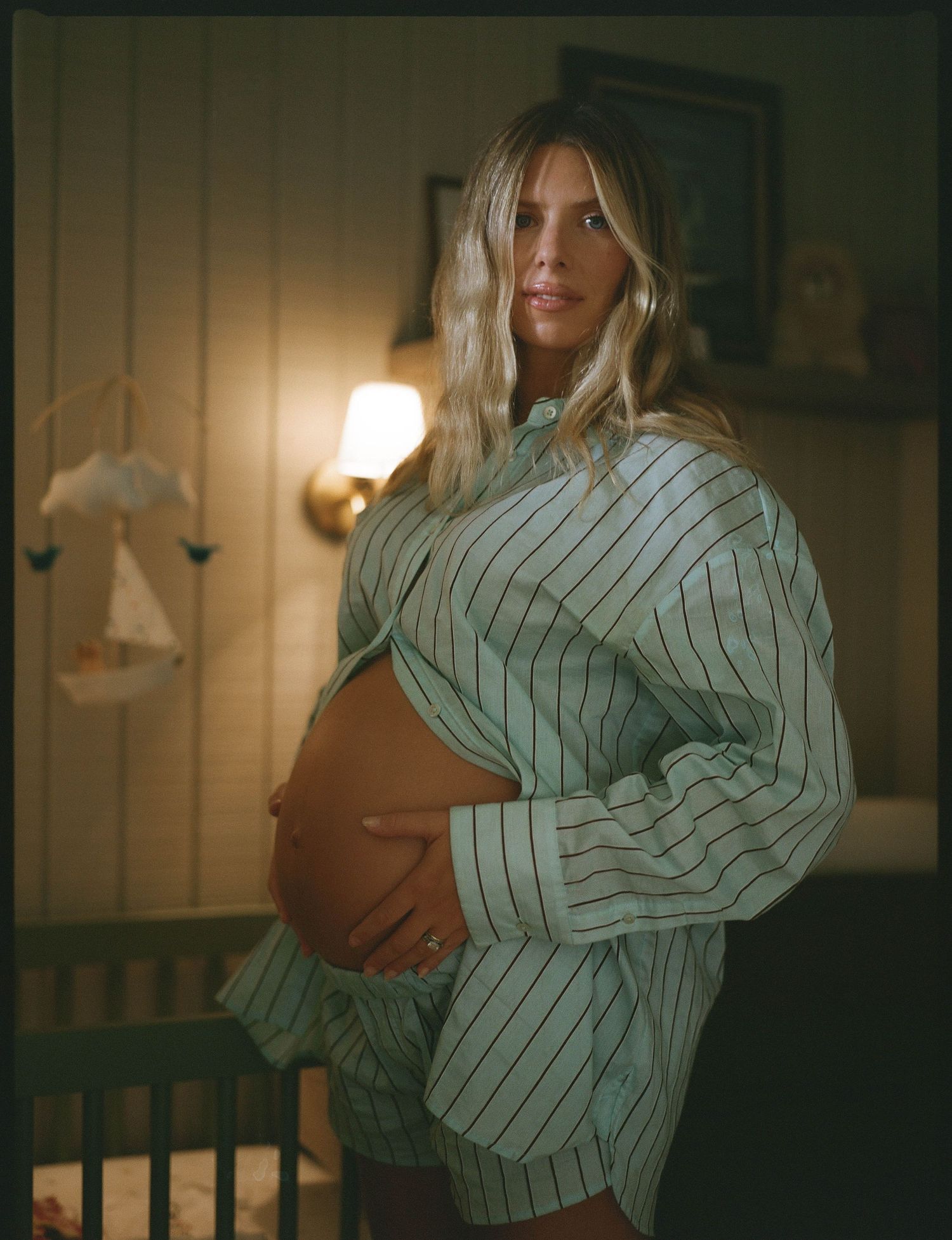 Pregnant woman in striped pajama set cradling baby bump in warmly lit nursery room.
