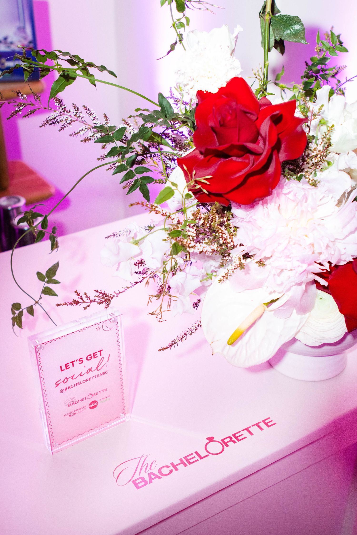 Table decoration with red roses and greenery against pink lighting and event signage.