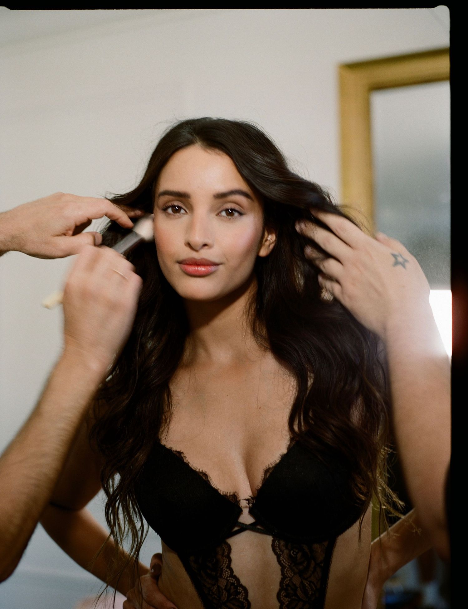 Woman with long wavy dark hair in black lace lingerie has makeup applied while stylists' hands fix her hair backstage.