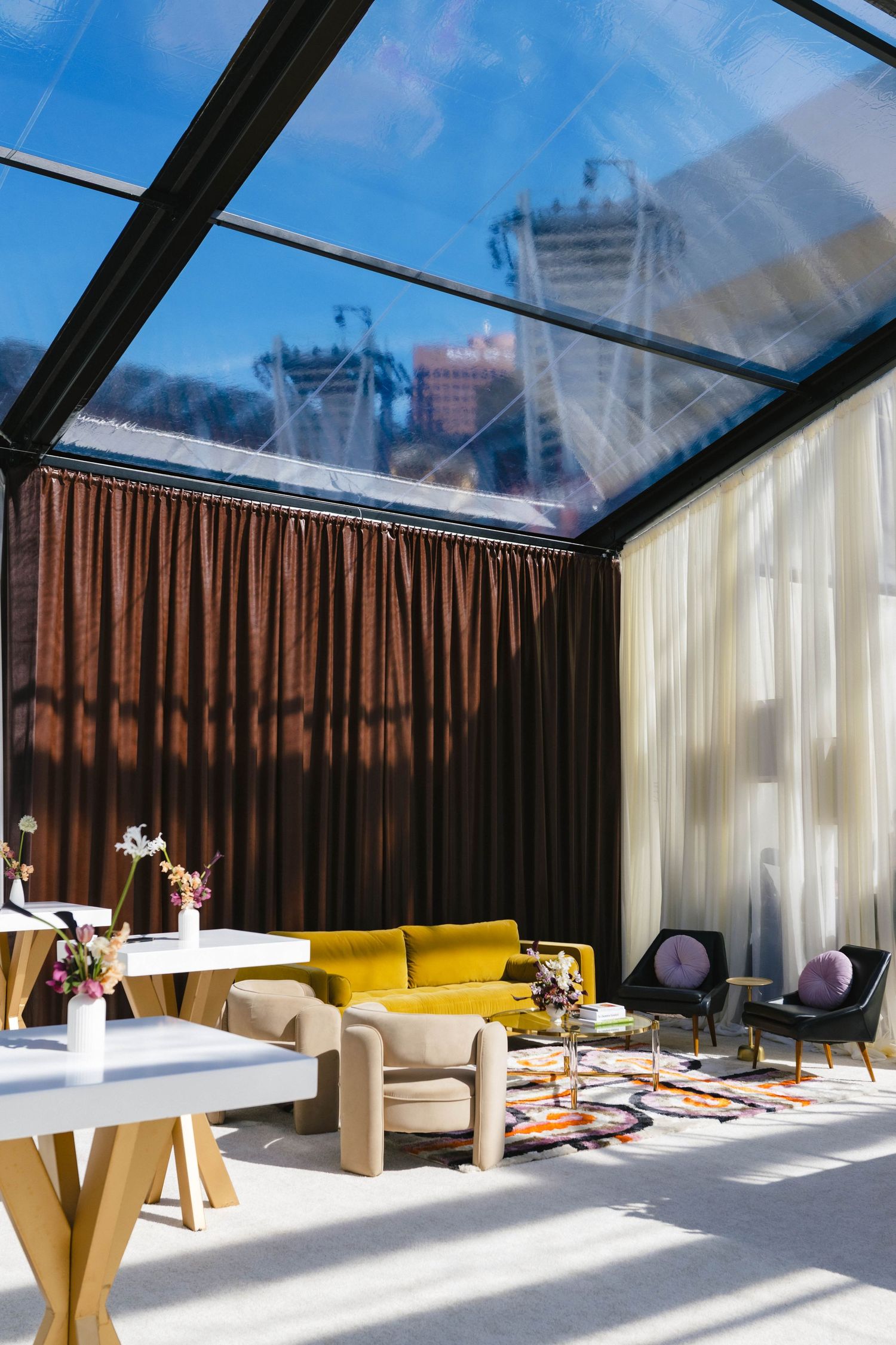 Modern glass-roofed lounge with velvet sofa, armchairs, colorful rug, and floor-to-ceiling curtains overlooking city skyline.