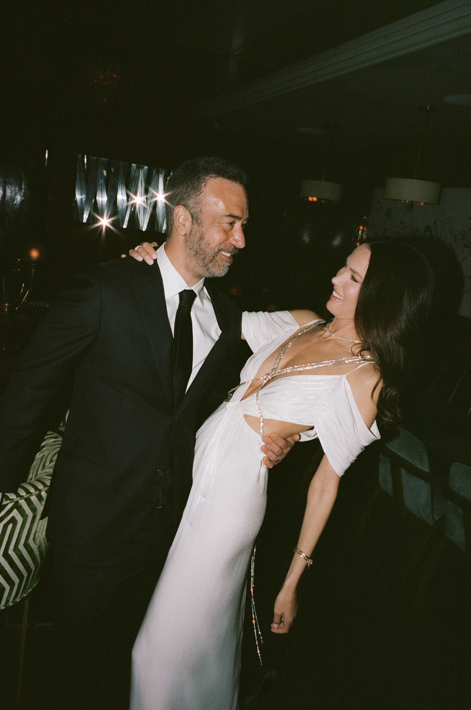 A man in a black suit and woman in a white gown share a joyful moment at an elegant evening event.