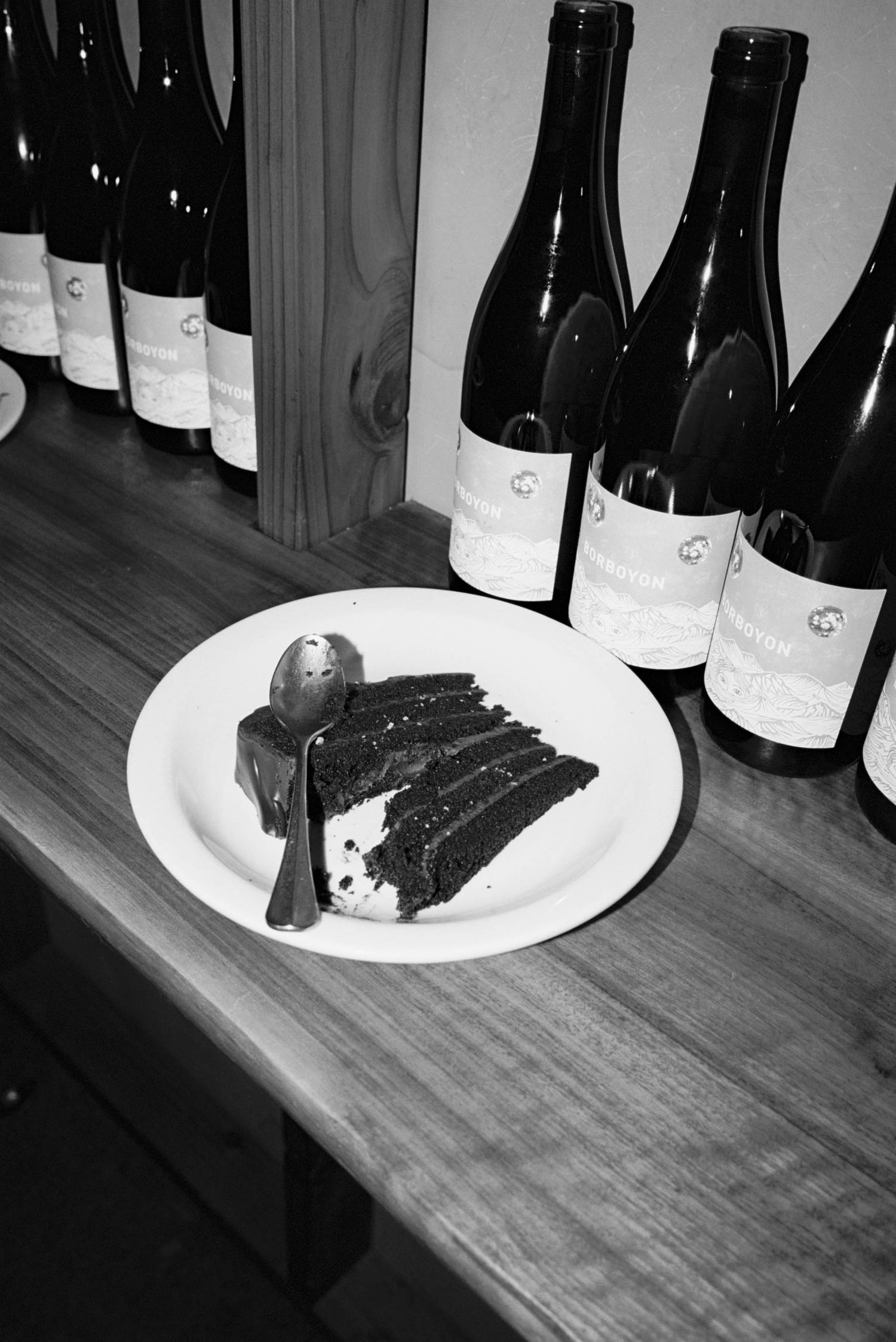 Black and white photo of chocolate cake slices with wine bottles in background.