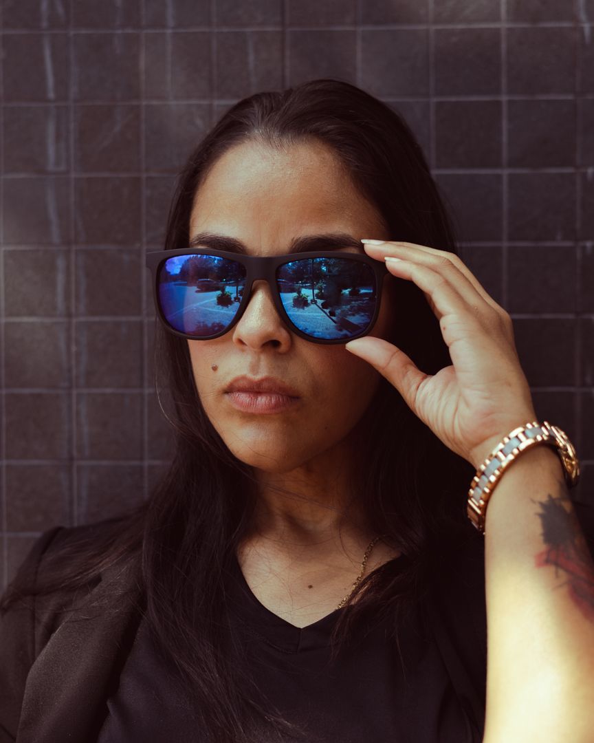 Person wearing reflective blue mirrored sunglasses and a black top poses against dark tiled wall.