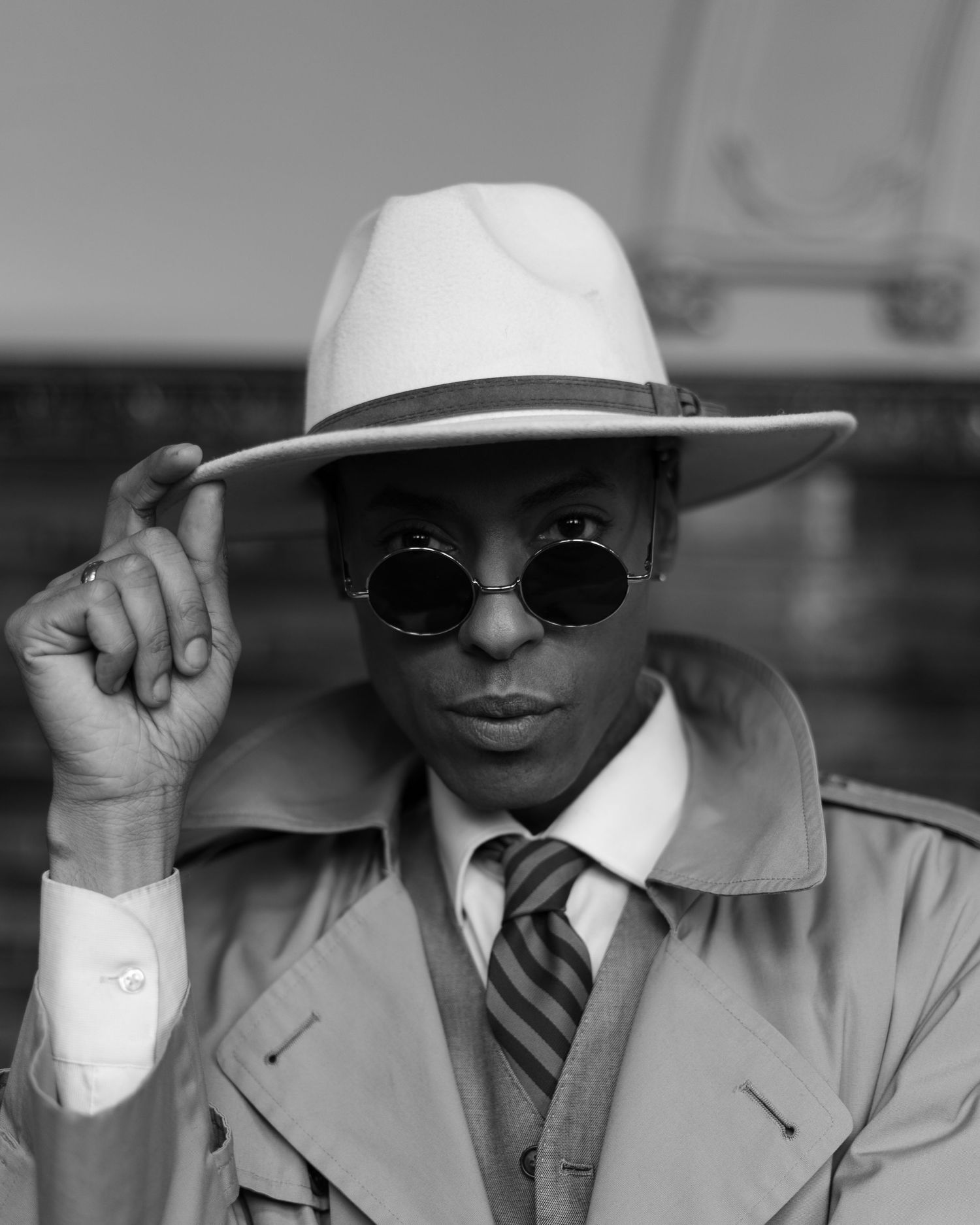 A person in a white suit and hat adjusts their brim while wearing sunglasses in black and white editorial photography.