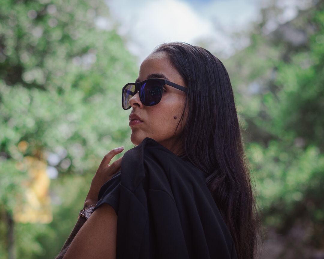 Woman with long dark hair wearing sunglasses poses in front of green foliage background.
