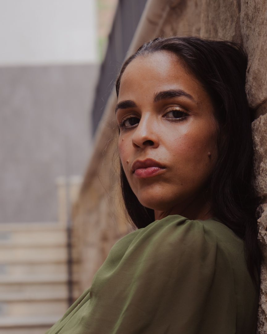 A young woman in a green top leans against a stone wall, gazing thoughtfully into the distance.