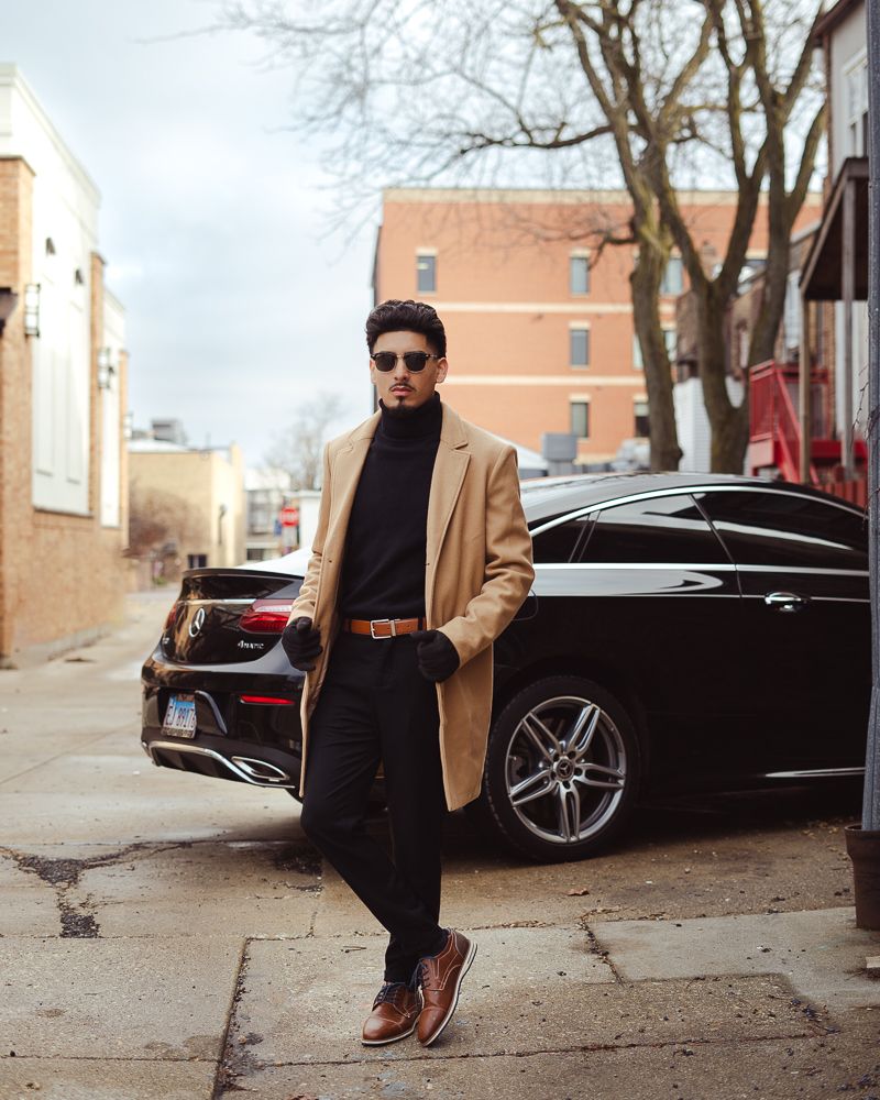 A person in a tan blazer and dark pants poses stylishly next to a sleek black luxury car on an urban street.