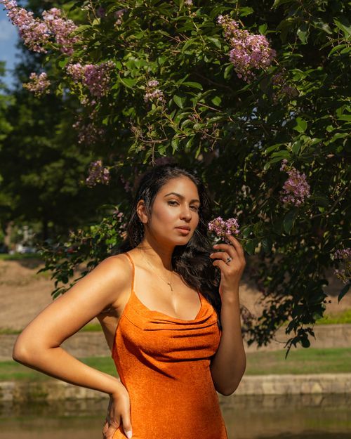A person in a bright orange dress poses elegantly near flowering trees in an outdoor setting.