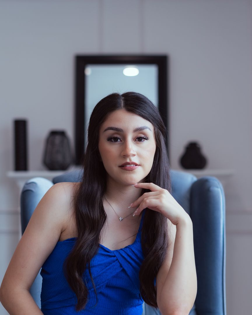Young woman in blue top sits in gray chair, long dark hair, hand on chin, elegant necklace, mirror in background.
