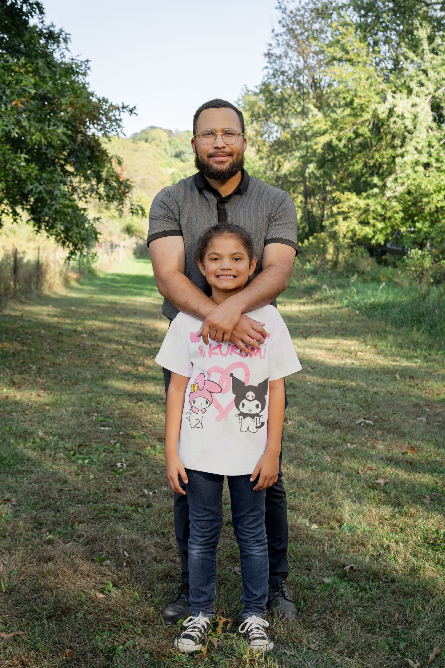 Parent and child in casual outfits pose together on grassy hillside.