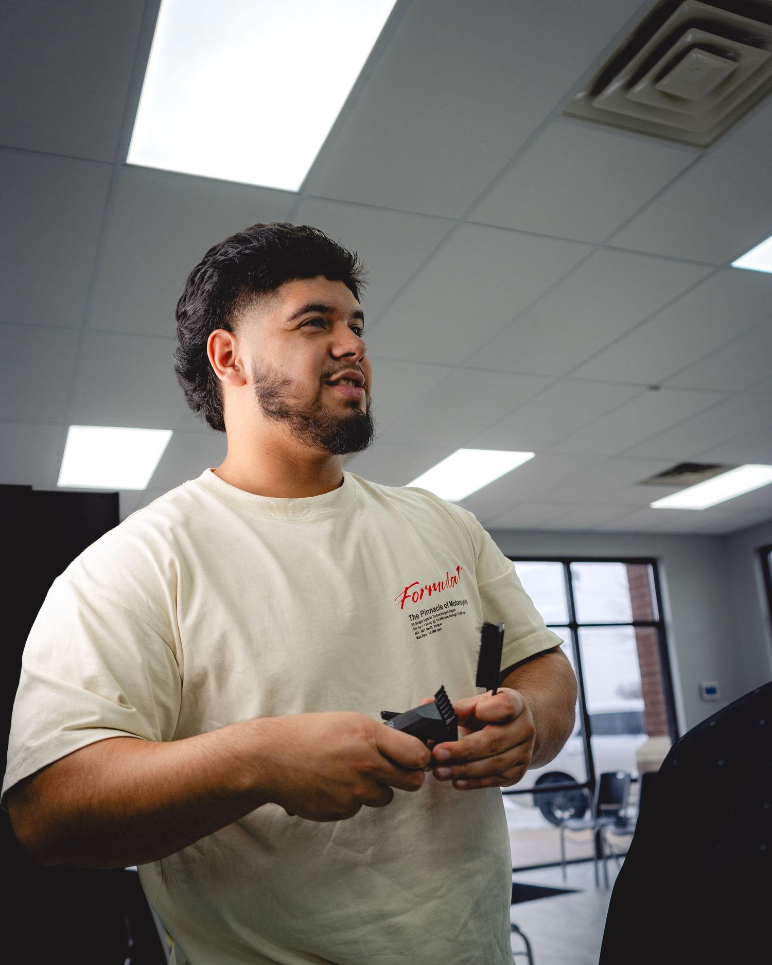 Person in a cream-colored t-shirt holding a device while standing in an office space with fluorescent lighting.