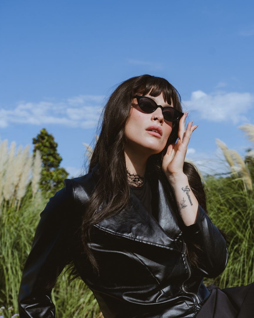 Woman in black leather jacket and sunglasses poses outdoors among tall grasses under a bright blue sky.