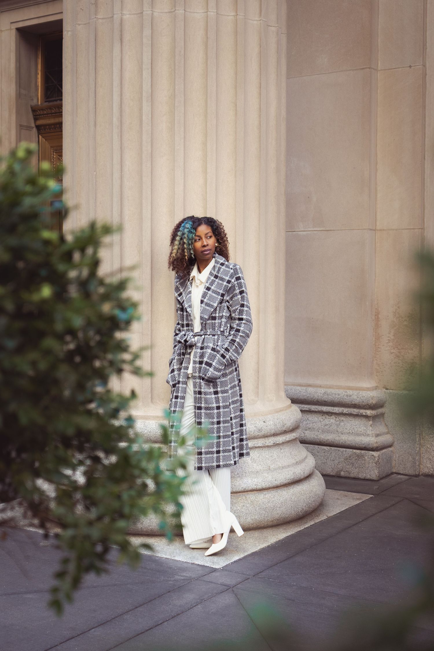Elegant couple posing against classical stone columns wearing plaid blazer and white dress.