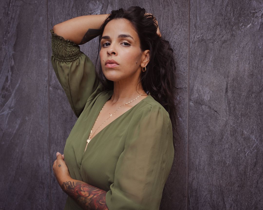 A woman in an olive green blouse poses confidently against a textured gray background.