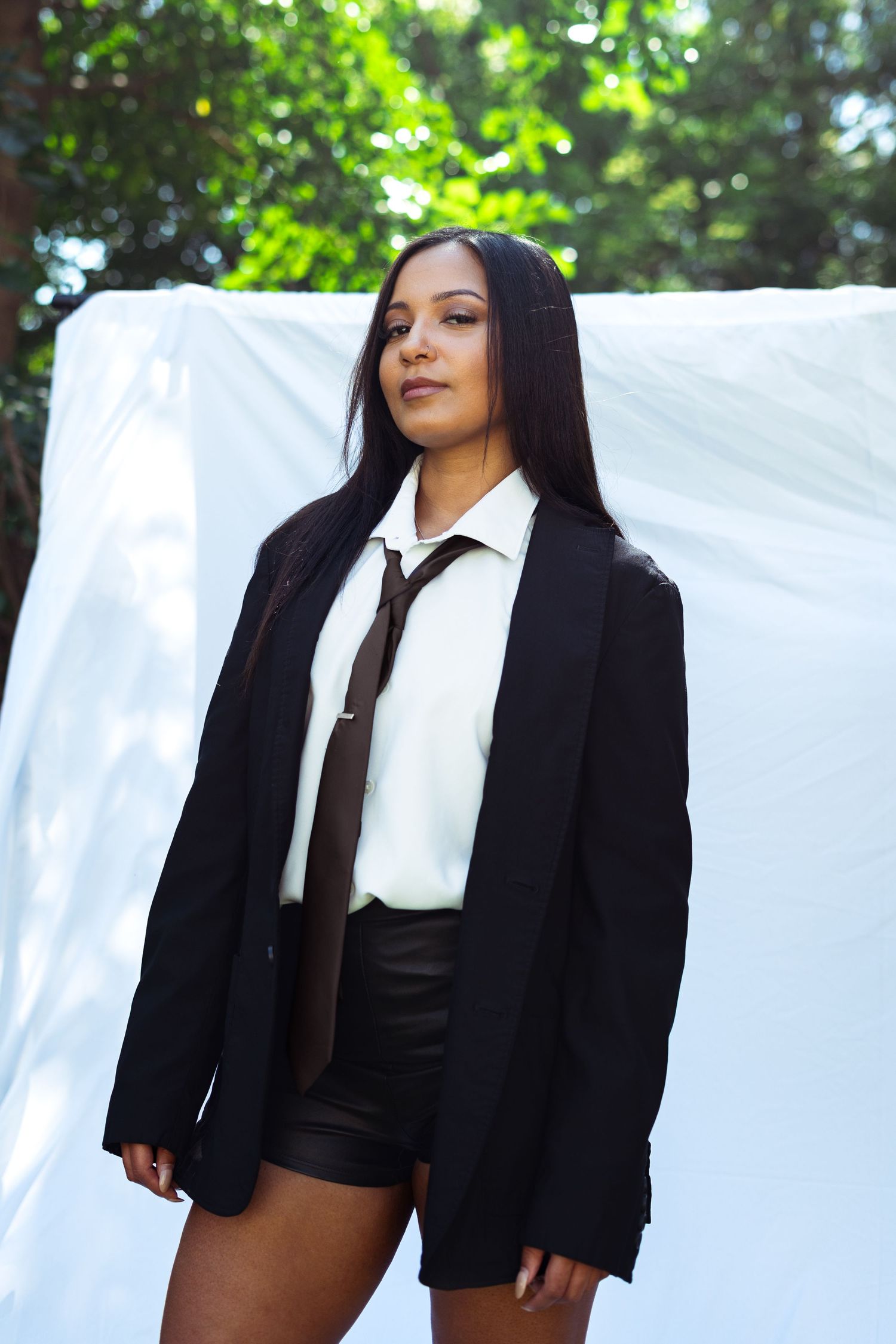 Editorial fashion model wearing a black blazer and tie poses outdoors against a white background.