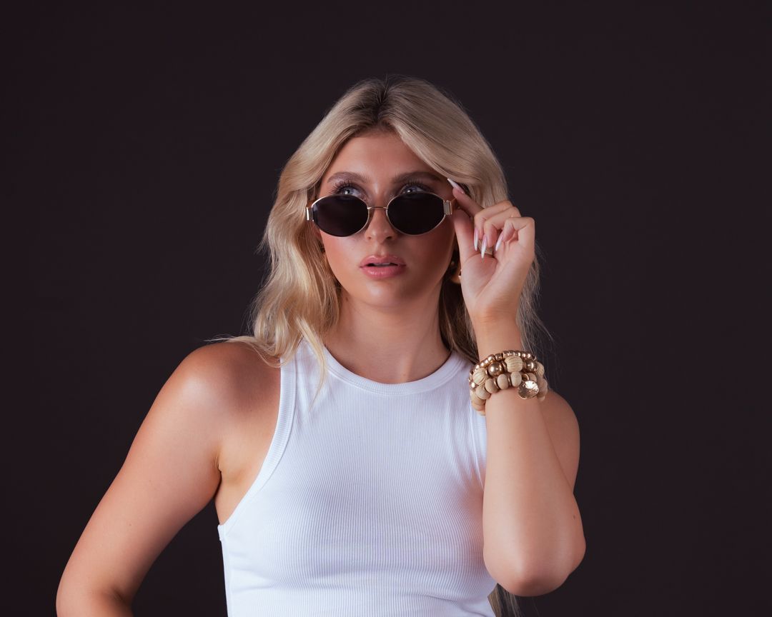 Blonde woman in white ribbed tank top wearing round sunglasses and gold bracelet posing against dark background.