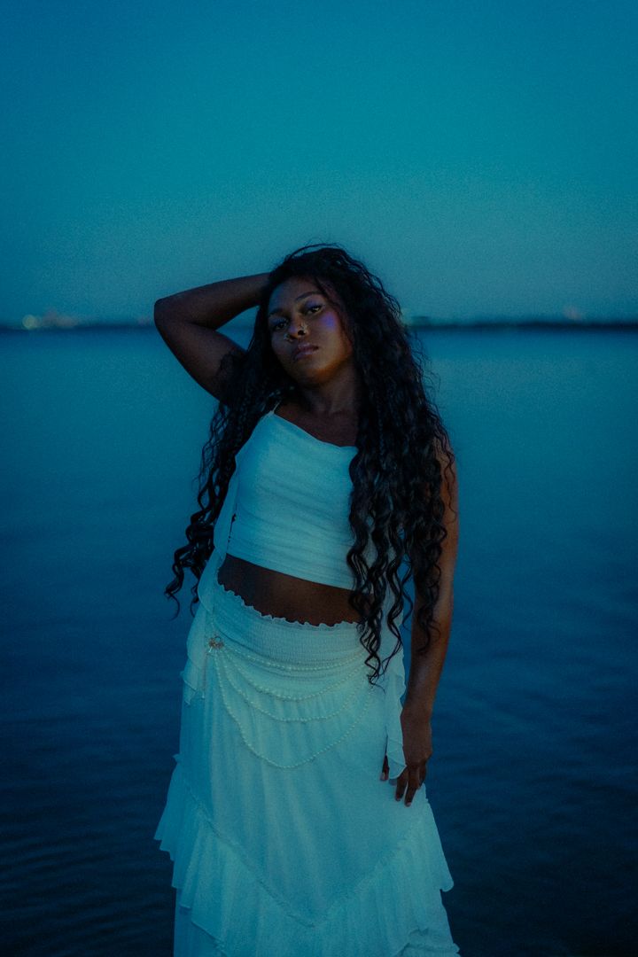 Artistic editorial photoshoot by water at dusk featuring white outfit against blue background.