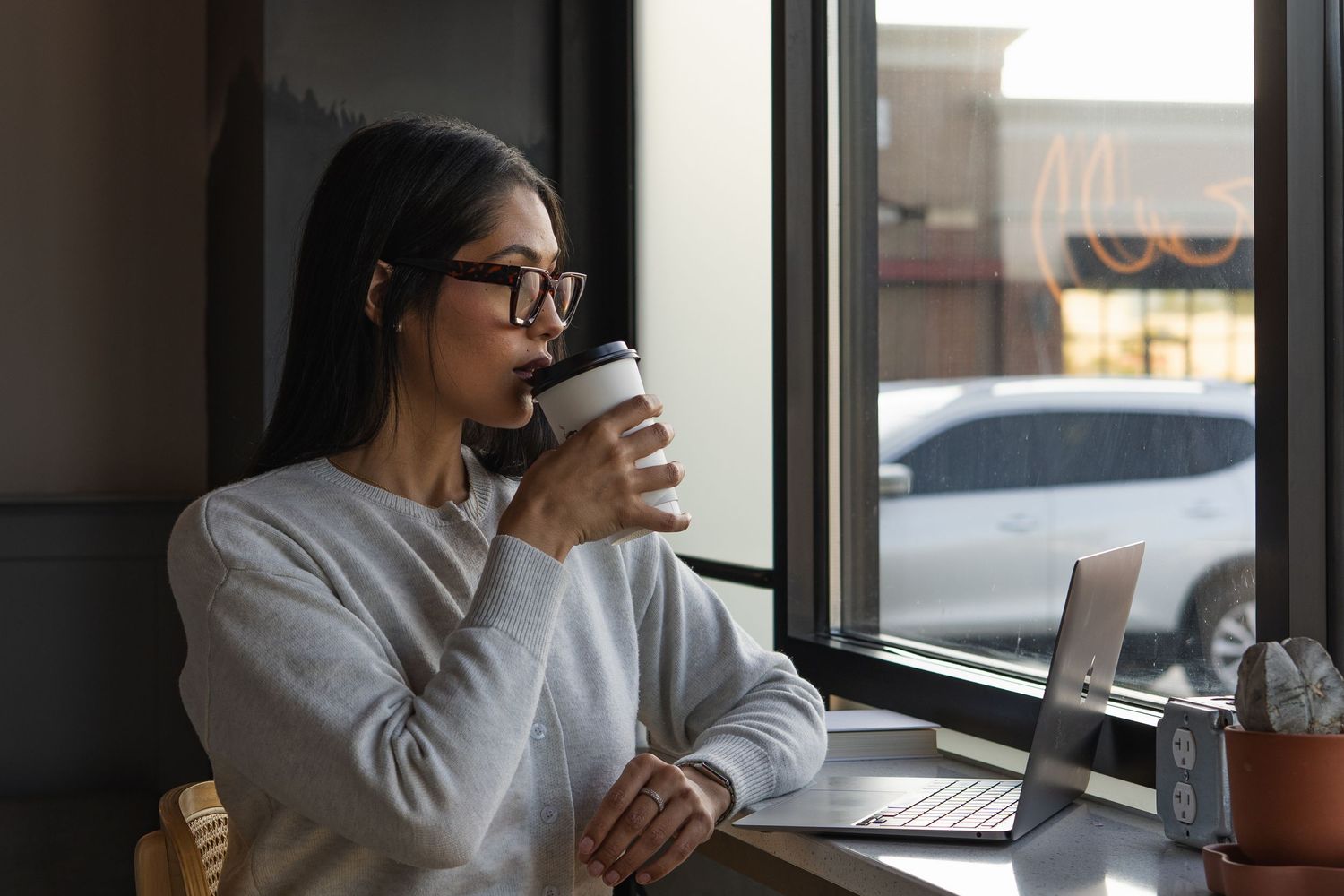 Young woman in glasses sipping coffee at cafe table with laptop near window.