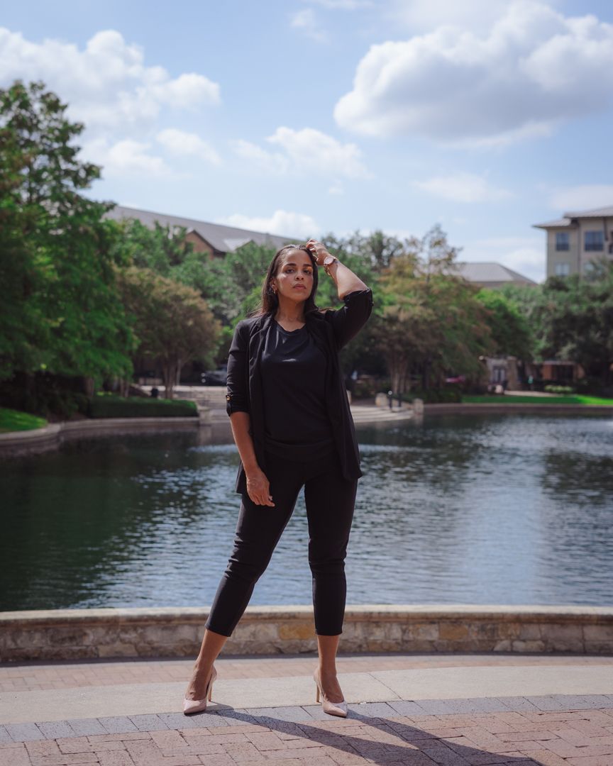 Person in all black outfit poses by a scenic pond with trees and buildings in the background on a sunny day.