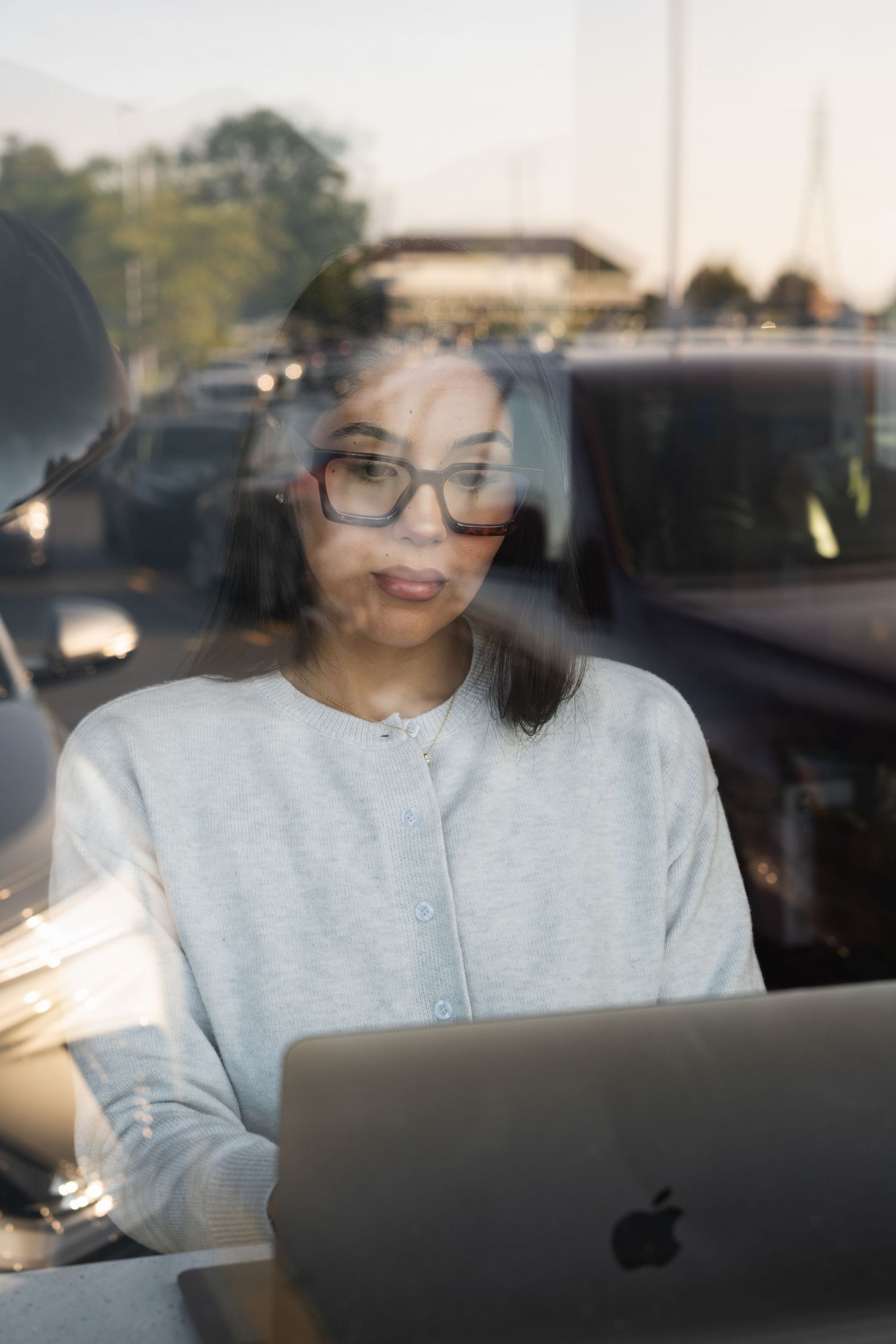 Woman with glasses working on Apple laptop, viewed through window with street reflections.