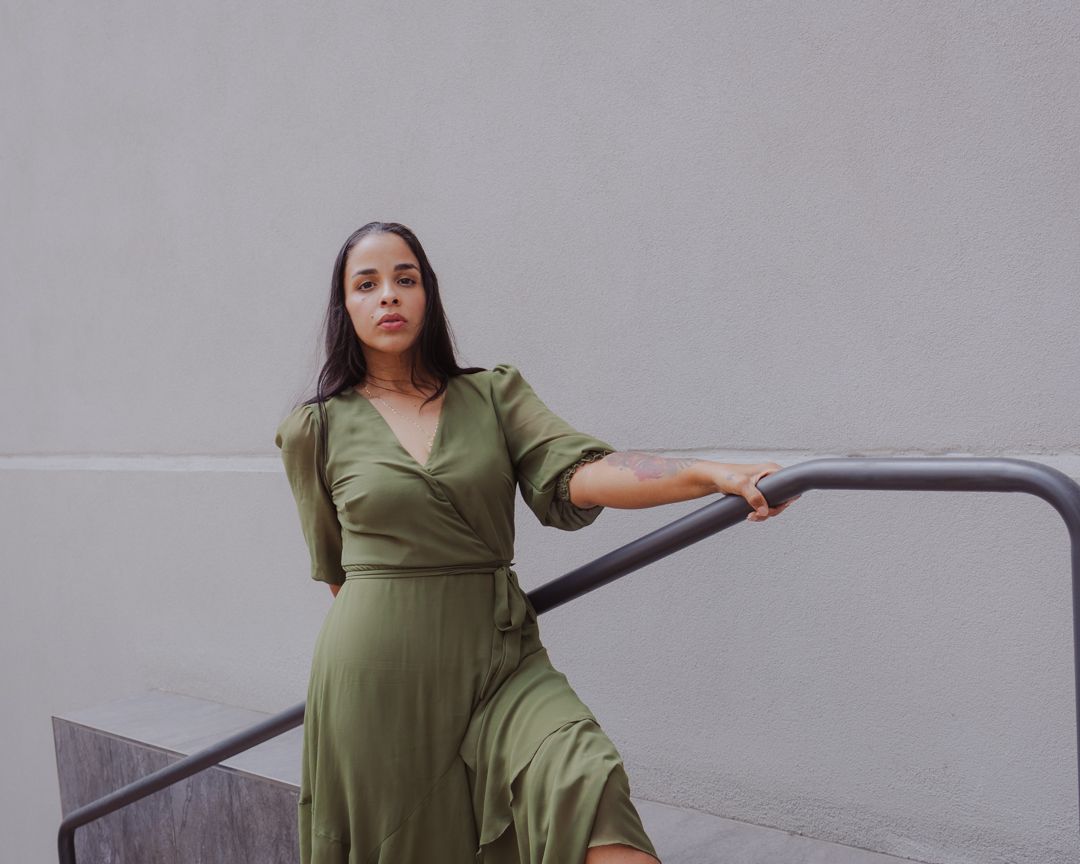 A woman in an olive green wrap dress poses confidently against a grey wall in various positions.
