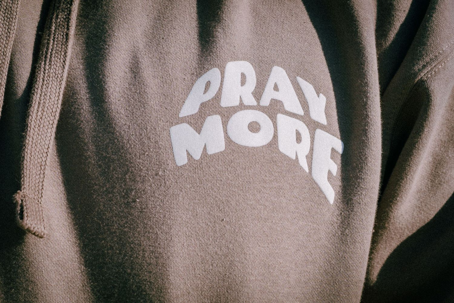 A brown hoodie sweatshirt with white text reading 'PRAY MORE' printed on the fabric.
