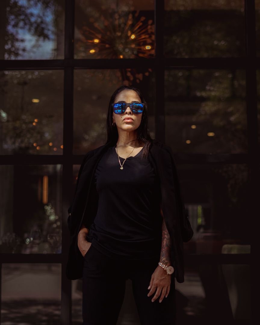 Stylish figure in black outfit and blue sunglasses posing against dark window backdrop.