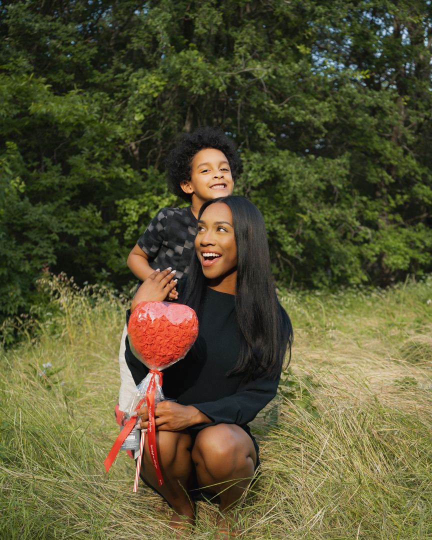 Person holding red heart-shaped lollipop while sitting in tall grass outdoors.