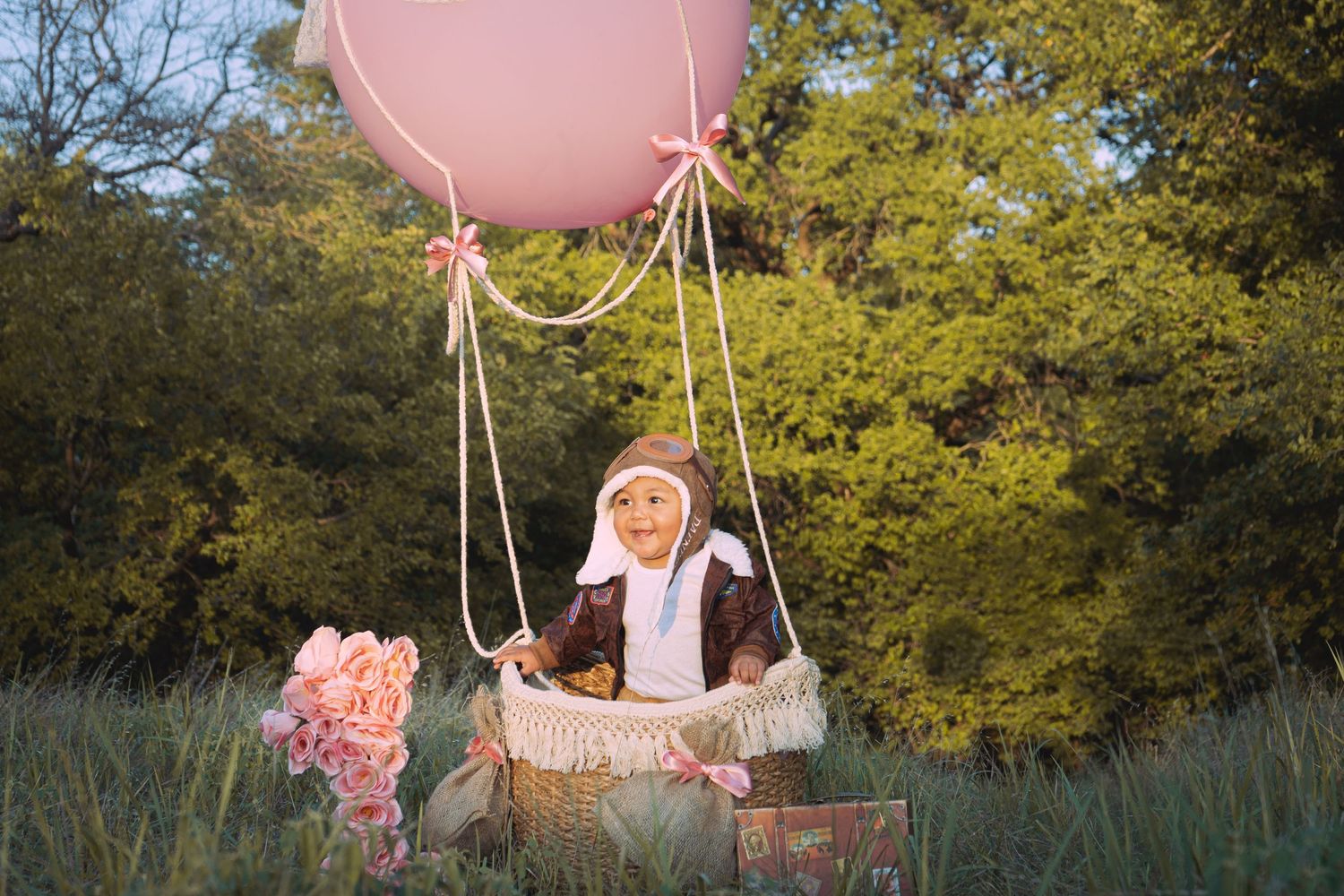 A whimsical outdoor scene with a wicker basket suspended by a large pink balloon in a natural setting at sunset.