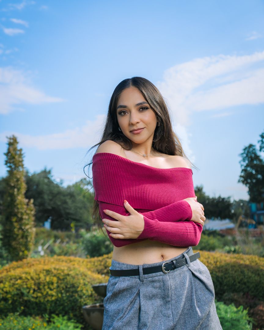 Young woman in pink off-shoulder ribbed top and gray trousers poses outdoors in a garden under a blue sky.