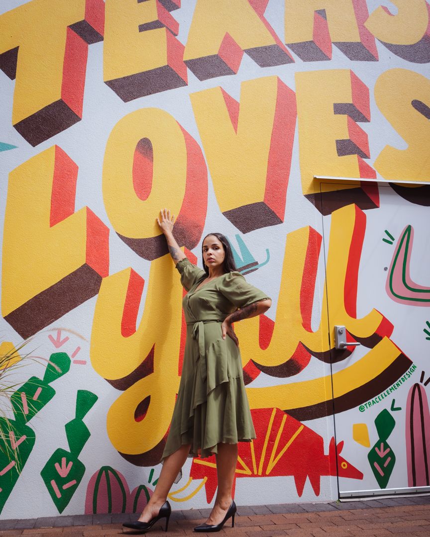 A person in an olive green dress poses against a vibrant wall mural featuring bold typography and geometric designs.