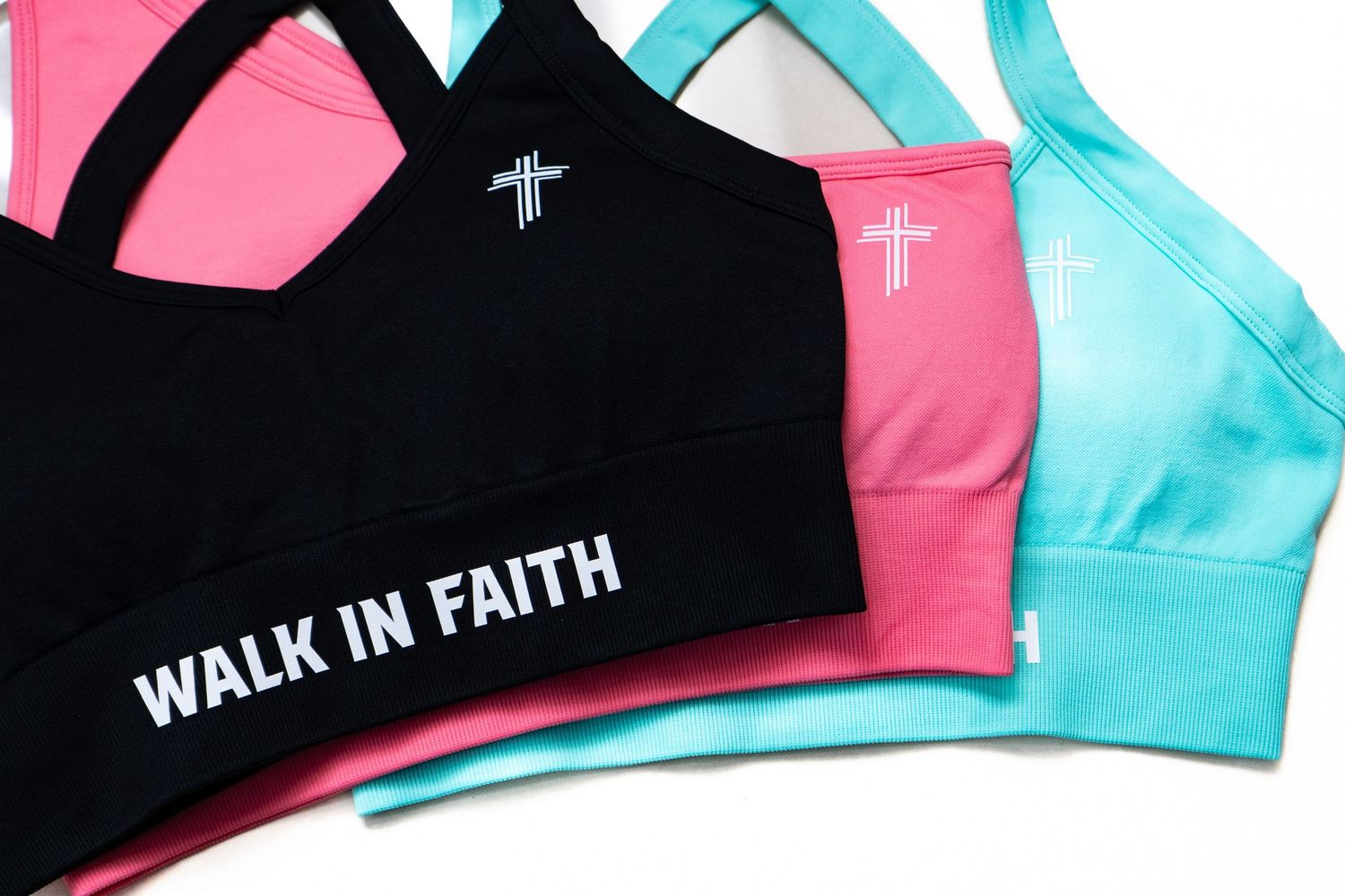 Three sports bras in black, pink, and teal with cross logos and Walk In Faith text, faith-inspired athletic wear.