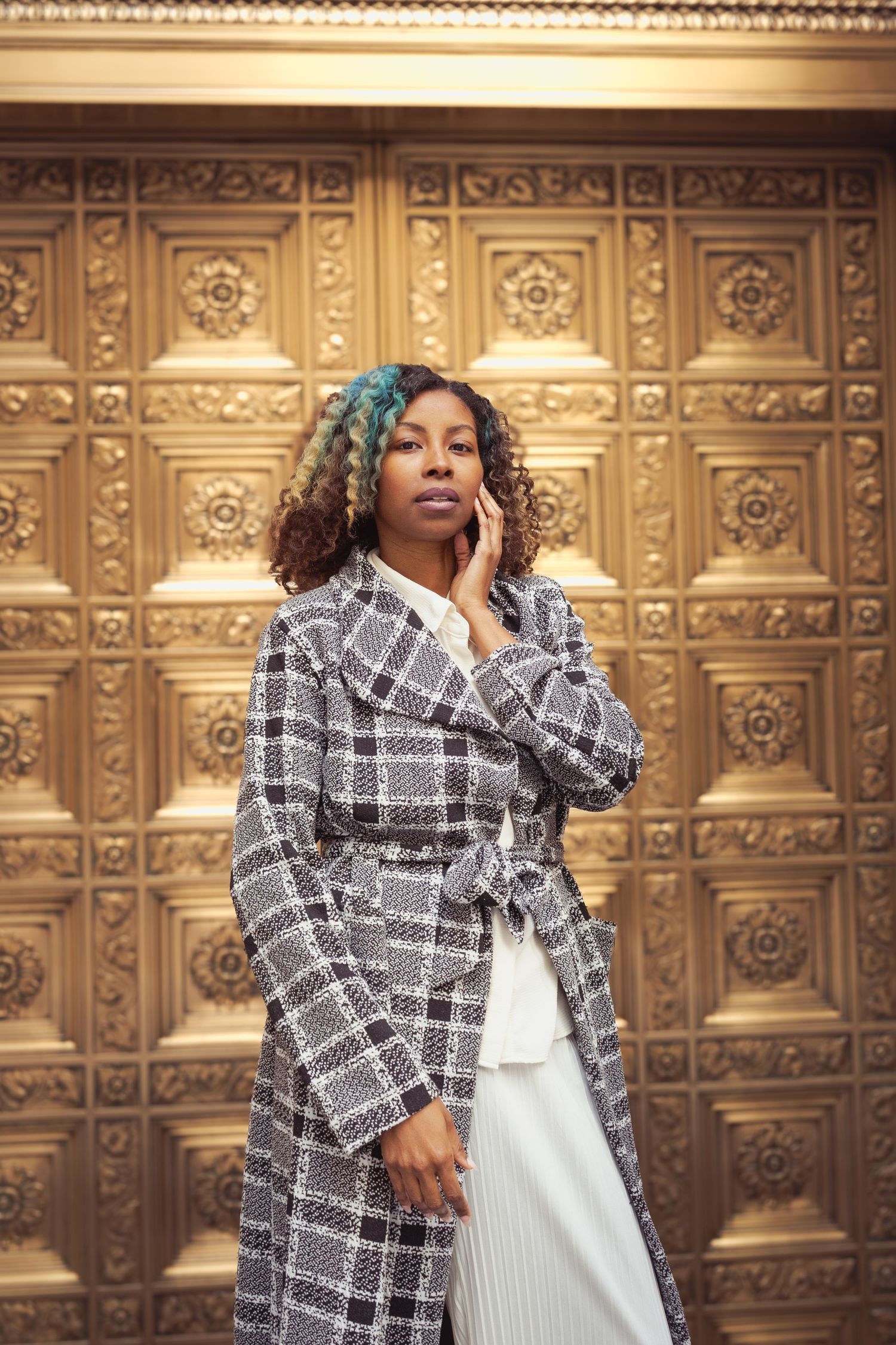 Model in plaid coat poses against ornate gold wall in fashion editorial photography.