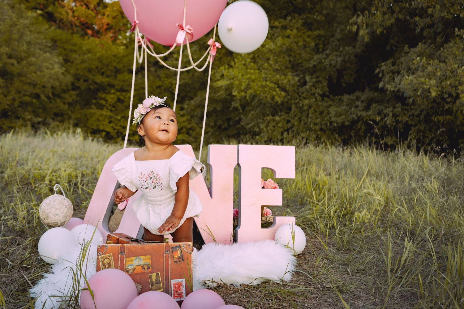 Pink birthday party setup in a meadow with vintage suitcase, tulle decorations and floating balloons for first birthday.