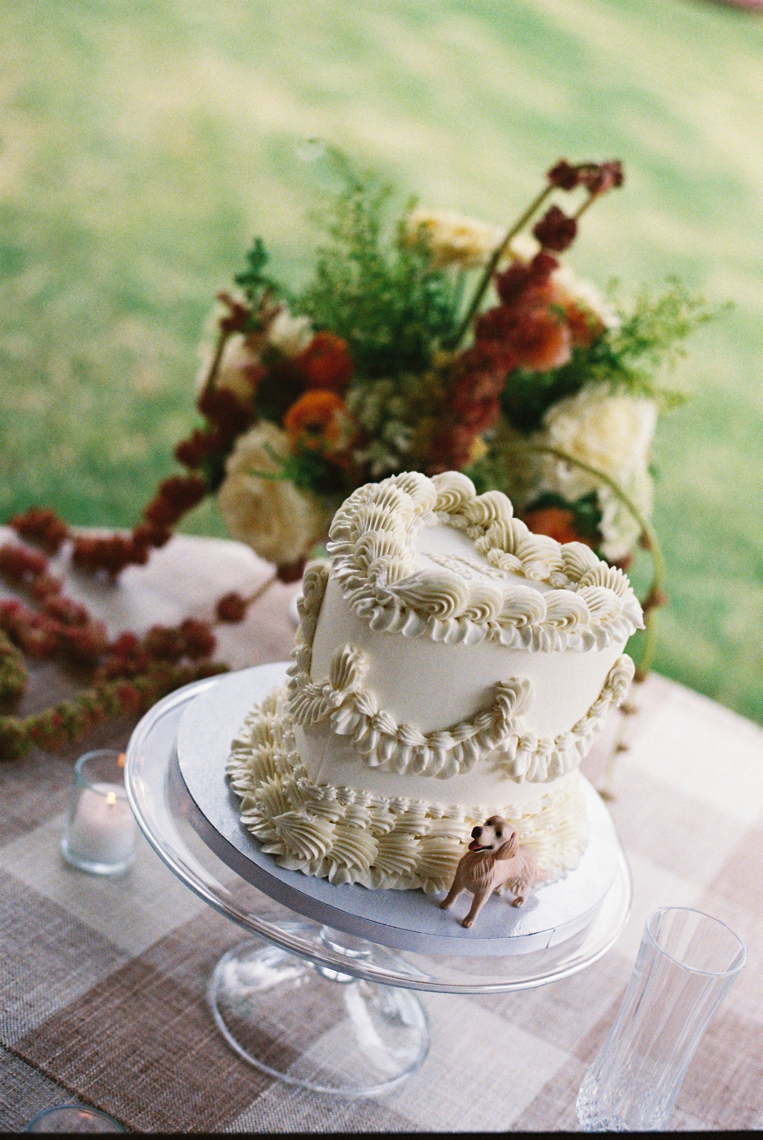 Two-tier white buttercream wedding cake with ruffled frosting on a glass stand, adorned with a small deer figurine.
