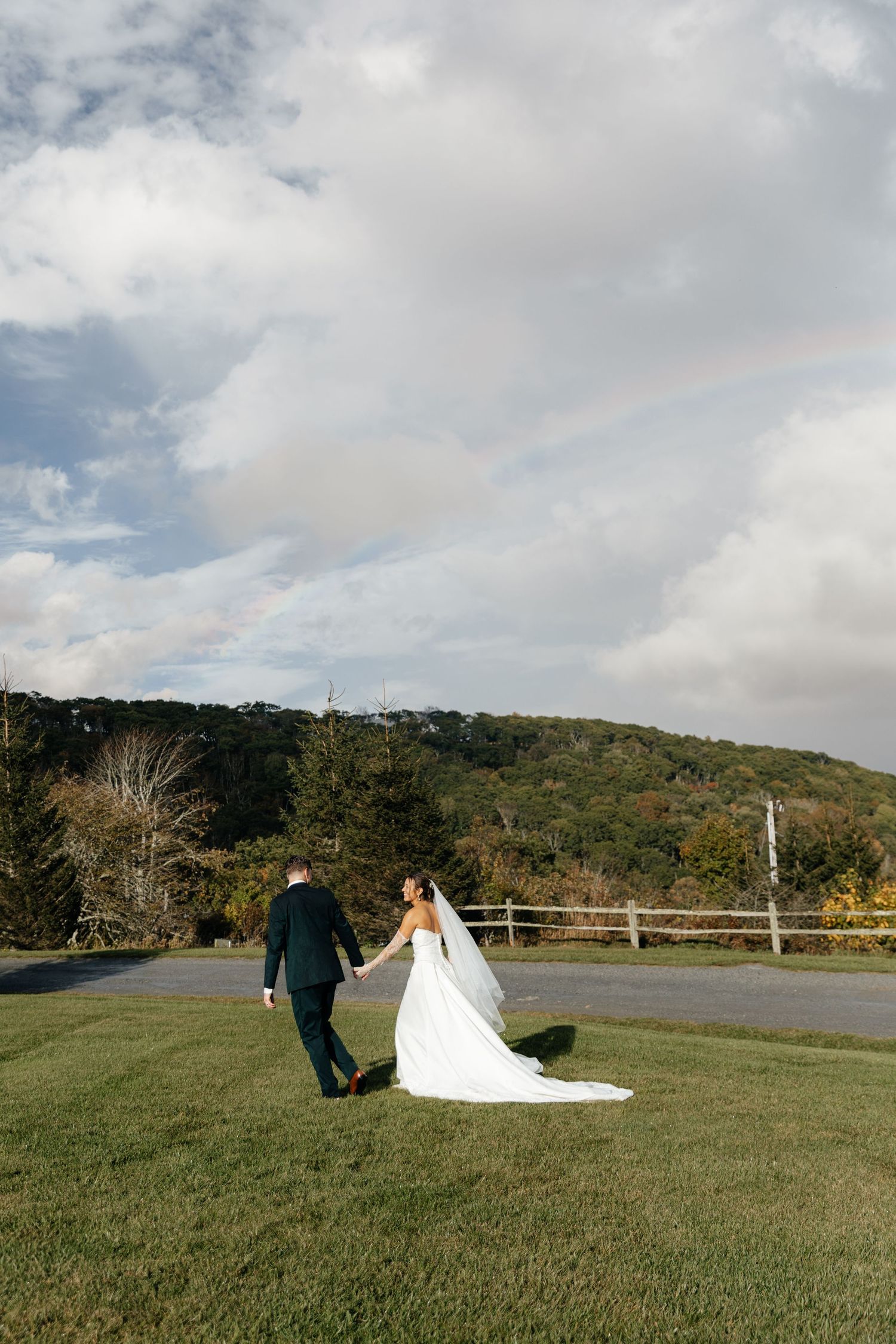 Bride and groom walk hand-in-hand across green lawn with rolling hills and dramatic cloudy sky backdrop.
