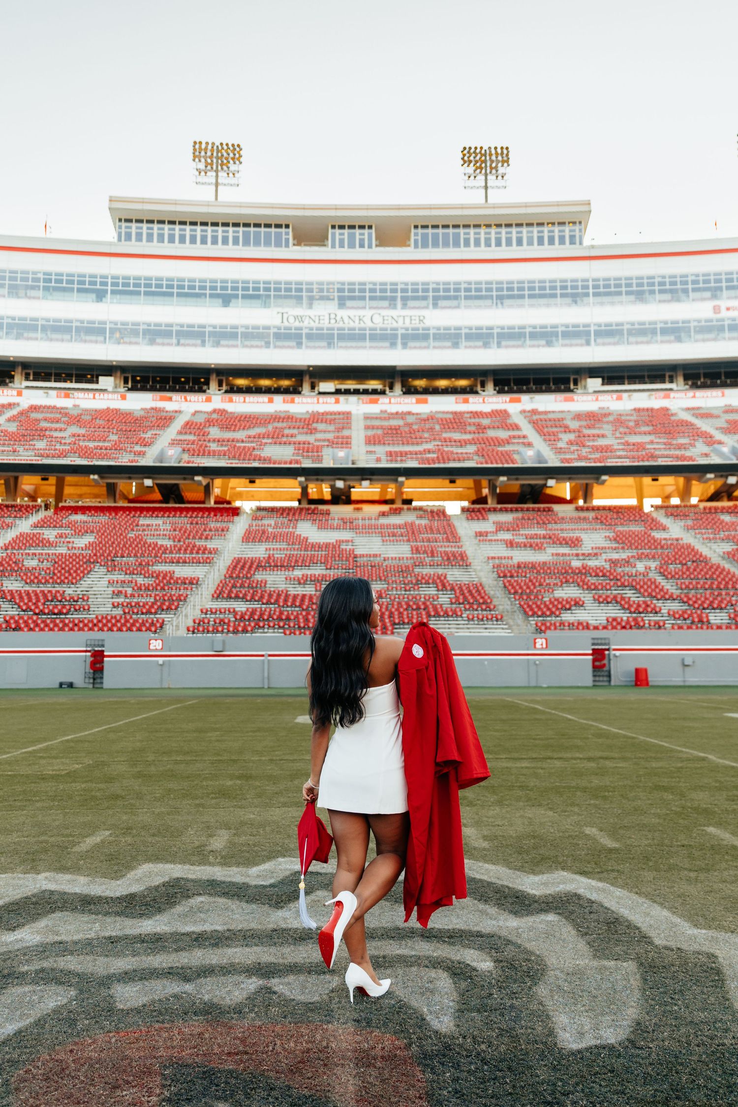 Graduate in white dress holds red gown on football field at empty stadium during graduation photoshoot.