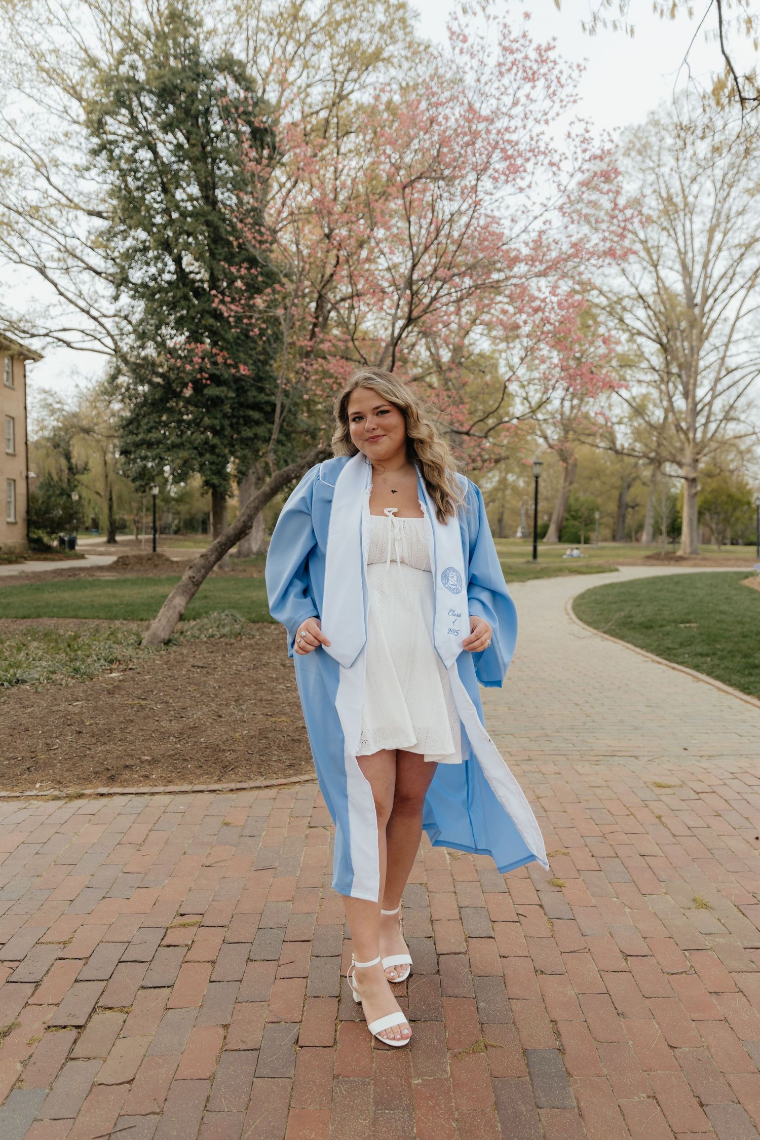 A young woman in a light blue graduation gown and white dress poses on a brick path surrounded by spring trees.
